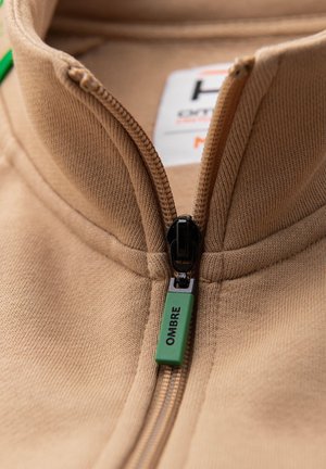 Beige lynlåsjakke med en grøn lynlåstrækker mærket "OMBRE" og en delvist synlig tøjmærkat inde i kraven.