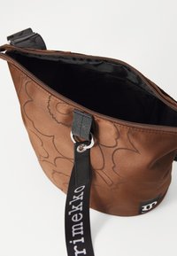 Brown fabric bucket bag with a floral embossed design, zip closure, black strap with logo, and a smooth inner lining. Silver hardware detail.