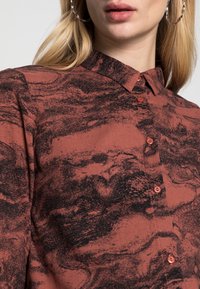 Camicia button-up in ruggine con motivo astratto nero, caratterizzata da un colletto classico e una texture particolare, accentuata da bottoni a contrasto.
