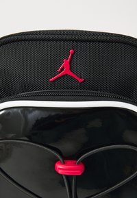 Borsa nera con finitura lucida, dettagli rossi e logo Jumpman. Presenta un materiale texturizzato e una chiusura con cordino elastico.