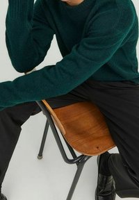 Tiffanyblå stickad tröja med hög hals, bärs med svarta byxor. Modellen sitter på en trästol och visar en avslappnad hållning och armposition.