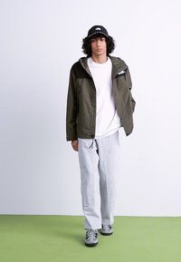 Wandeljack in olijfgroen met zwarte accenten, wit T-shirt, lichtgrijze cargo broek en grijze sneakers, staand op groene vloeren.