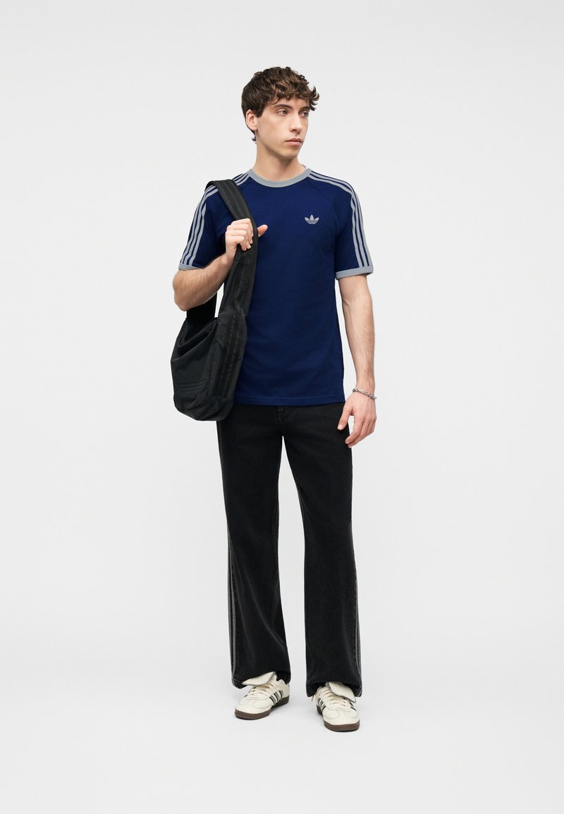 Giovane uomo con t-shirt Adidas blu navy, pantaloni neri, sneakers bianche, che tiene una borsa a tracolla nera, in piedi contro uno sfondo chiaro e uniforme.