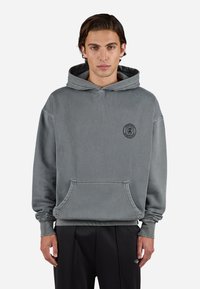 Grijze hoodie met een voorzak, verstelbare trekkoordcapuchon en een zwart rond logo op de borst. Het materiaal lijkt zacht en textuur te hebben.