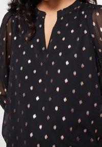 Blouse noire avec un col en V et des manches transparentes, décorée de motifs de points métalliques en or rose sur tout le tissu.