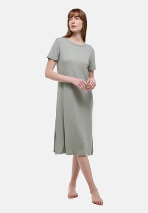 Femme aux cheveux bruns lisses portant une robe midi ample à manches courtes de couleur gris clair, debout pieds nus sur un fond blanc.