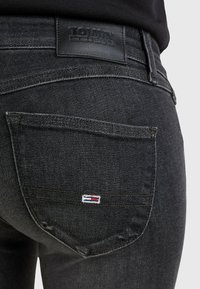 Vaqueros de mezclilla negra con un parche de cuero de la marca Tommy Jeans en la cintura y un pequeño logo bordado en el bolsillo trasero.