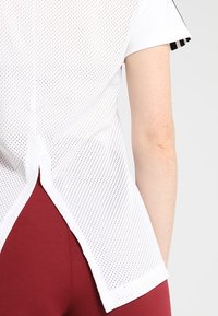 Vit nättröja med korta ärmar, med en delad avslutning och svarta detaljer. Materialet är ventilerande och lätt.