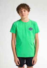 Groene katoenen T-shirt met korte mouwen, ronde hals en een klein blauw diamanten logo op de borst. In combinatie met zwarte shorts.