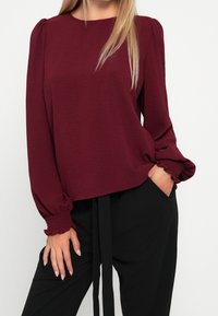 Burgundy långärmad blus med samlade ärmar och texturerad tyg. Matchad med högmidjade svarta byxor med knytmidja.