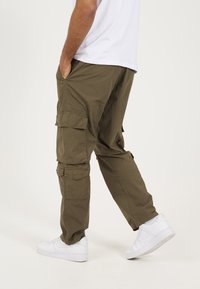 Ελαιώδεις πράσινοι cargo παντελόνια με πολλαπλές τσέπες, άνετη εφαρμογή και ελαφριά υφή. Συνδυασμένα με λευκά αθλητικά παπούτσια για αντίθεση.