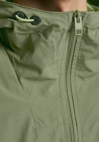 Giacca verde con zip e colletto alto, dotata di cordoncino, texture liscia e dettagli di cucitura sottili.