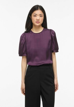 Sirgete tumedate juustega naine, kes kannab läikivat lilla pliiatsvarrukatega pluusi ja kõrge vöökohaga musti pükse, valge tausta ees.