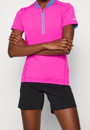 Henkilö, jolla on käsivarret ristissä, pukeutuneena kirkkaan vaaleanpunaiseen lyhythihaiseen vetoketjulliseen paitaan ja mustiin shortseihin valkoista taustaa vasten.