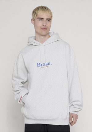 Jeune homme aux cheveux blonds courts portant un sweat à capuche gris clair avec l'inscription « Broke. » en bleu sur la poitrine, debout devant un fond uni.