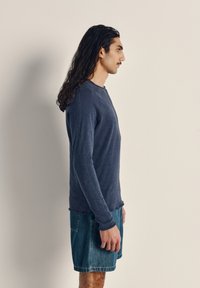 Ilga rankovė, tamsiai mėlyna medvilninė marškinė su apvaliu kaklu, derinama su džinsiniais šortais. Lygus tekstūra ir kasdieniškas pjūvis.