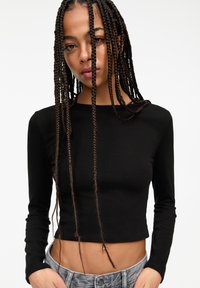 Schwarzes langärmliges Crop-Top aus dehnbarem Material. Figurbetonter Schnitt mit rundem Ausschnitt. Kombiniert mit hochgeschnittenen Jeans.