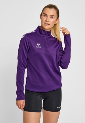 Top sportivo viola con zip e maniche lunghe, colletto alto e strisce bianche sulle spalle. Indossato con pantaloni corti neri; presenta il logo sul petto.
