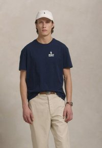 Fiatal férfi sötétkék Polo Ralph Lauren pólót, bézs nadrágot, fehér sapkát és karórát visel, egy sima háttér előtt áll.