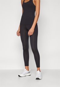 Svart tettsittende tank topp kombinert med svarte leggings, begge laget av elastisk materiale. Hvite sportssko har et mesh-design og snøring.