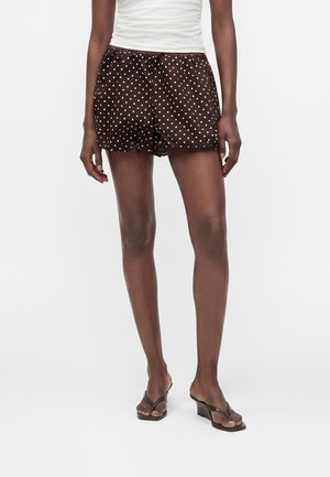 Femme portant un short marron à pois, un top blanc sans manches et des sandales compensées noires, debout devant un fond blanc uni.