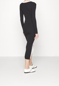 Persona che indossa un vestito midi nero a maniche lunghe con costine e uno spacco laterale, abbinato a sandali bianchi flat su uno sfondo semplice.
