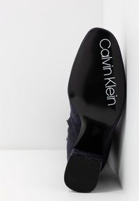 Semelle d'une chaussure à talons hauts noire Calvin Klein montrant le logo et le talon texturé sur une surface blanche contre un mur blanc.