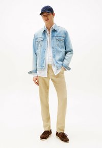 Denim jakna v svetlo modri barvi, z dvema žepoma na prsih in gumbnim zapiranjem, v plasteh nad belim črtastim srajcem, v kombinaciji z bež hlačami in rjavimi čevlji.