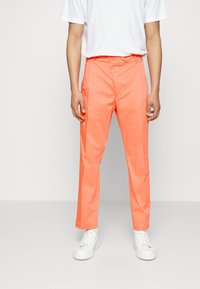Helle orange Hose mit glatter Textur, ausgestattet mit einem Knopfverschluss und Seitentaschen, kombiniert mit weißen Turnschuhen und einem schlichten weißen T-Shirt.