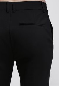 Svarta byxor gjorda av ett glansigt, lätt tyg. Funktioner inkluderar en bakficka med en smal öppning och en skräddarsydd passform.