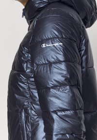 Person med en glänsande svart puffjacka med Champion-logotyp broderad på överarmen, med dragkedja och huva.