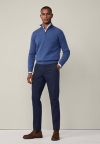 Niebieski dzianinowy sweter z zamkiem przy kołnierzu, zestawiony z granatowymi spodniami slim-fit. Całość dopełniają brązowe zamszowe botki za kostkę. Widoczne szczegóły w fakturze materiału.