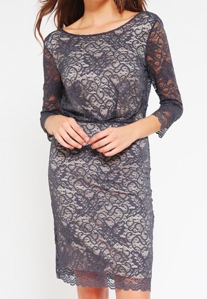 Frau trägt ein knielanges, figurbetontes graues Spitzenkleid mit Dreiviertelärmeln und einem wellenförmigen Saum über einem hautfarbenen Futter.