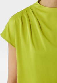 Limonkowa bluzka z krótkimi rękawami, subtelnymi fałdami na ramionach oraz teksturowaną powierzchnią z delikatnymi paskami. Gładkie wykończenie materiału.