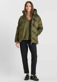 Olijfgroen pufferjack met een capuchon, ritsluiting en trekkoorden, gecombineerd met een donkergroen bovenstuk en zwart gestreepte broek. Zwarte schoenen.