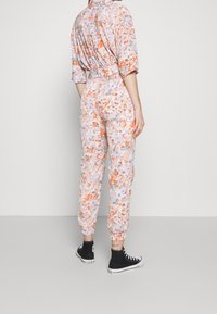 Mono floral en rosa pastel, con mangas cortas y cintura elástica. Decorado con acentos en naranja y azul verdoso; pantalones tapered con puños.