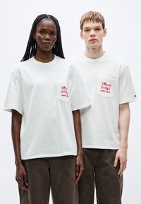 Camisetas blancas de manga corta con logotipos rojos en el bolsillo. Cuello redondo, corte holgado. Combinadas con pantalones marrones de tela texturizada.