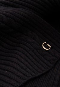Bufanda negra de punto acanalado con un acento de logo circular dorado. La textura es suave, con un patrón de cable prominente en toda la superficie.