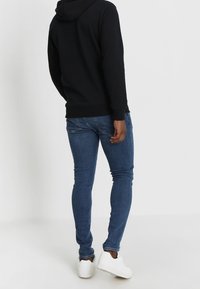 Sudadera con capucha negra, jeans skinny azules y zapatillas blancas. Tejido suave, diseño ajustado, costuras visibles y jeans de largo hasta el tobillo.