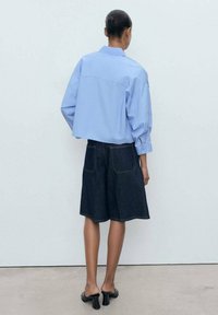 Lichtblauwe, geknipte knoopdas met pofmouwen, gecombineerd met donkere denim shorts. Eenvoudig ontwerp, achteraanzicht, gedragen met zwarte hoge hakken.