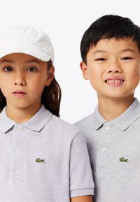 Grå pikétröjor med korta ärmar; knappar vid kragen; Lacoste-logotyp på vänster bröst. Ett barn bär en vit keps.
