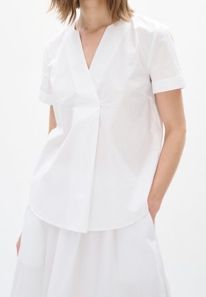 Kvinde iført en hvid V-hals bluse med korte ærmer og hvid nederdel, stående med den ene hånd i lommen mod en ensfarvet hvid baggrund.