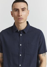 Camisa de manga corta en color marino, fabricada en tejido texturizado, con cierre de botones en la parte delantera, cuello clásico y un bolsillo en el pecho.