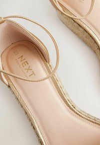 Sandales à talons dorées avec une semelle en espadrille texturée, un dessus en cuir lisse et un design à fines lanières. Nom de marque "NEXT" gravé à l'intérieur.