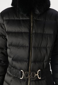 Zwarte gewatteerde jas met een bontkraag, gouden rits en een gestructureerde tailleband voorzien van een metalen riemgesp met de letter "M."