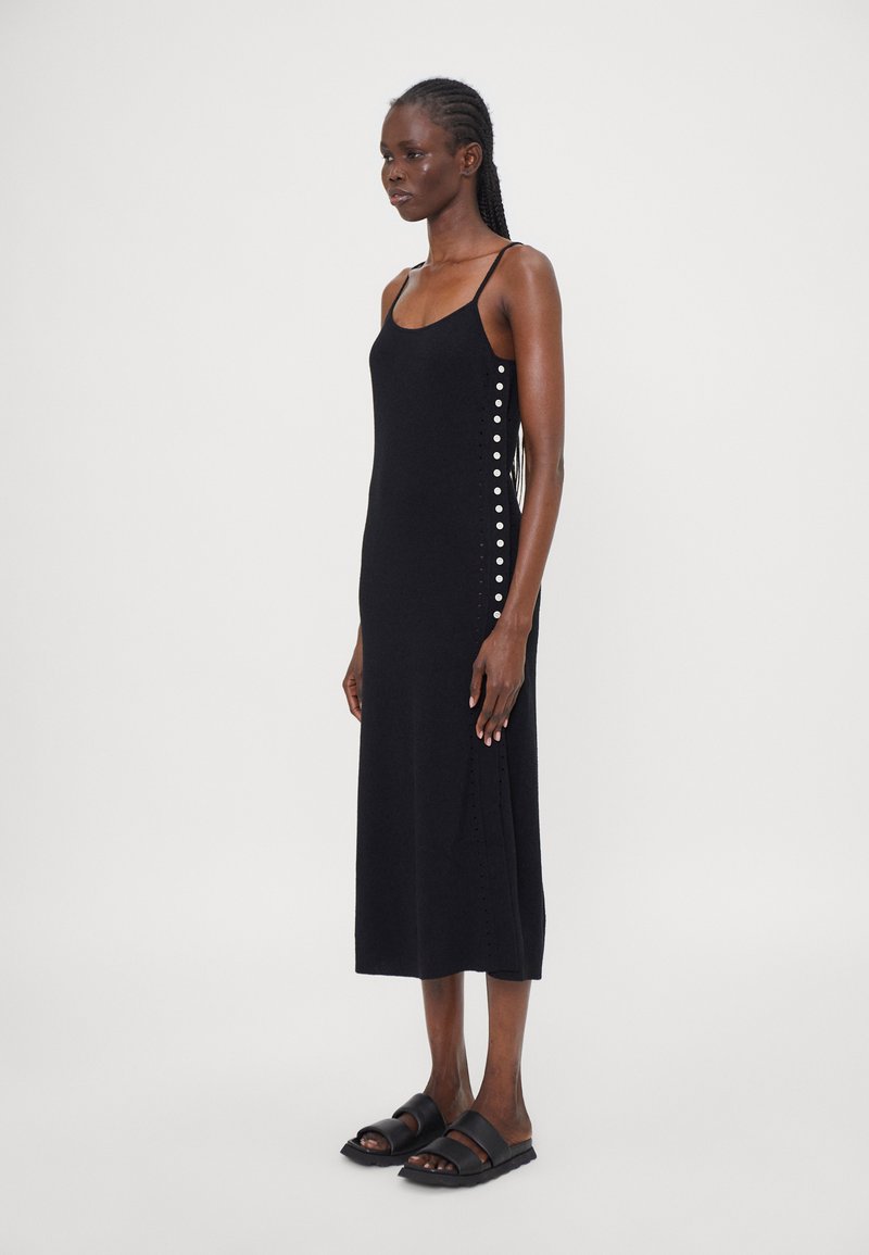 Schwarzes gestricktes Maxikleid mit dünnen Trägern, seitlichem Knopfdetail und einem leichten Schlitz. Kombiniert mit schwarzen Riemchensandalen.
