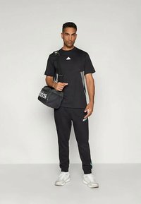 Juodas sportinis kostiumas su trumpomis rankovėmis marškinėliais su baltu logotipu ir juostelėmis, derančiomis kelnėmis ir juoda sportine krepšiu su baltu prekės ženklo ženklu.