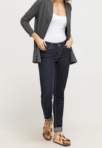 Granatowe dżinsy o dopasowanym kroju, zestawione z białym topem na ramiączkach i szarym kardiganem. Brązowe sandały z klamrami dopełniają stylizację.