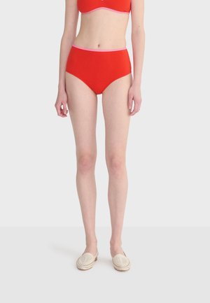 Kobieta w czerwonej bieliźnie z wysokim stanem z różowym obszyciem oraz kremowych wsuwanych butach plecionych, stojąca na tle jasnego, jednolitego tła.