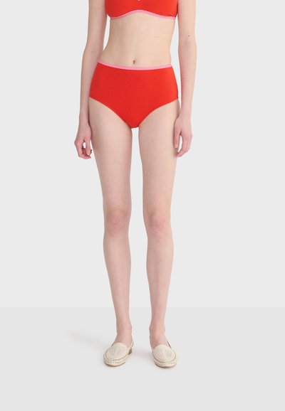 Femme portant une culotte taille haute rouge avec un bord rose et des chaussures à enfiler en crème tissée, debout devant un fond clair uni.
